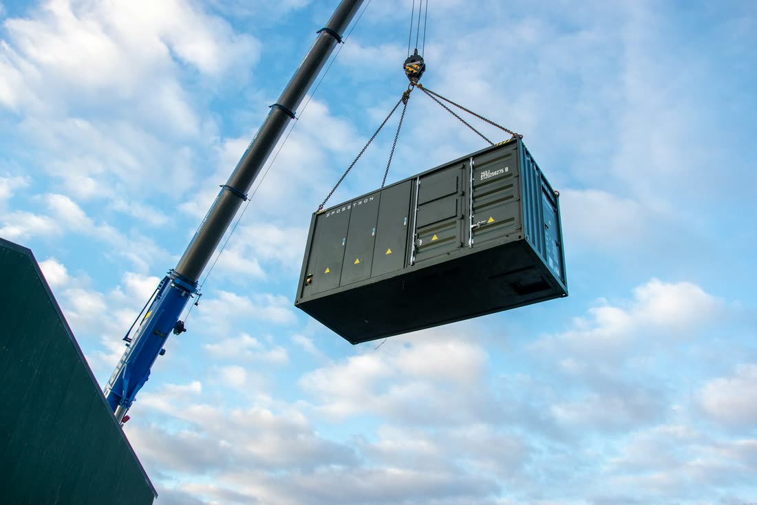 A crane lifts a dark green battery storage container against a pale blue sky